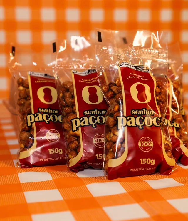 Três pacotes vermelhos de Amendoim Caramelizado Senhor Paçoca de 150 g sobre toalha xadrez laranja